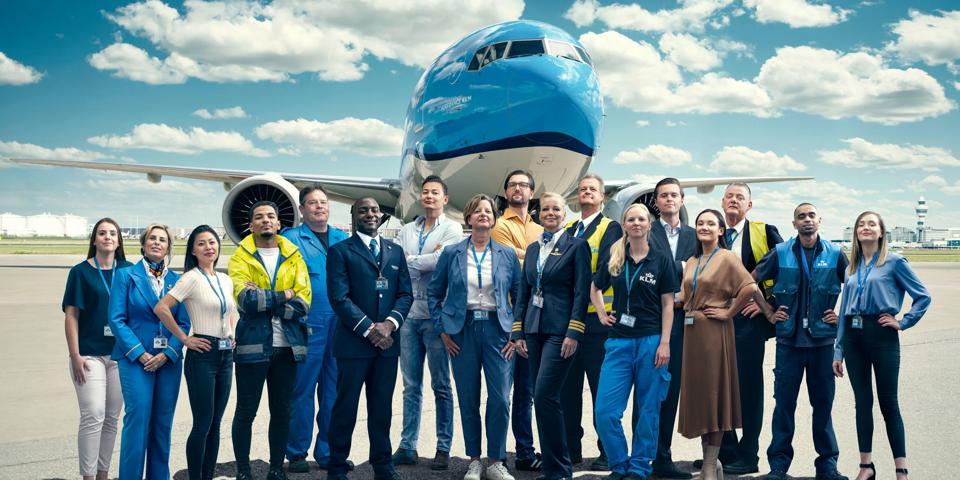 Diverse KLM staff from all divisions proudly posing on the tarmac in front of a large airplane, smiling in their uniforms under a partly cloudy sky.
