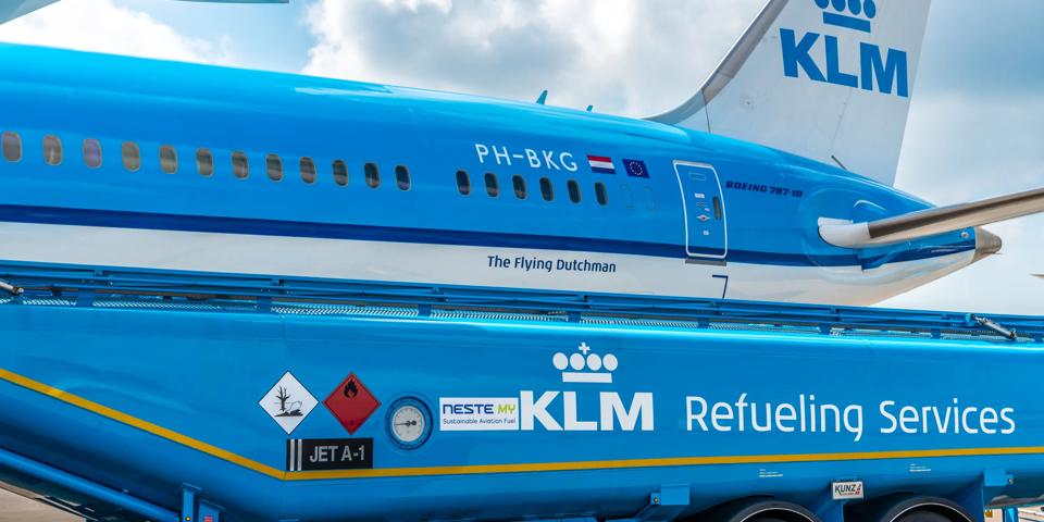A KLM plane is being refuelled with SAF, an alternative to fossil aviation fuel. In the foreground, a KLM-branded refuelling truck with a Neste logo is supplying fuel to the aircraft. The sky is partly cloudy.
