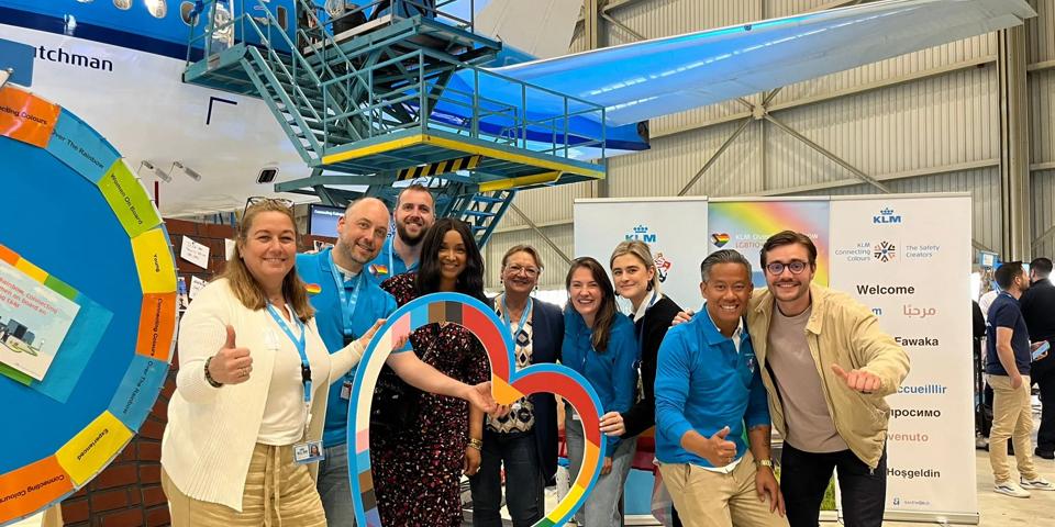 Group of people holding a rainbow shaped heart near a KLM plane.