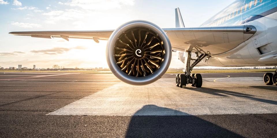 Low-angle view of a KLM airplane on the runway, with a large jet engine in the foreground, landing gear beneath the wing, and warm sunrise light reflecting on the aircraft.